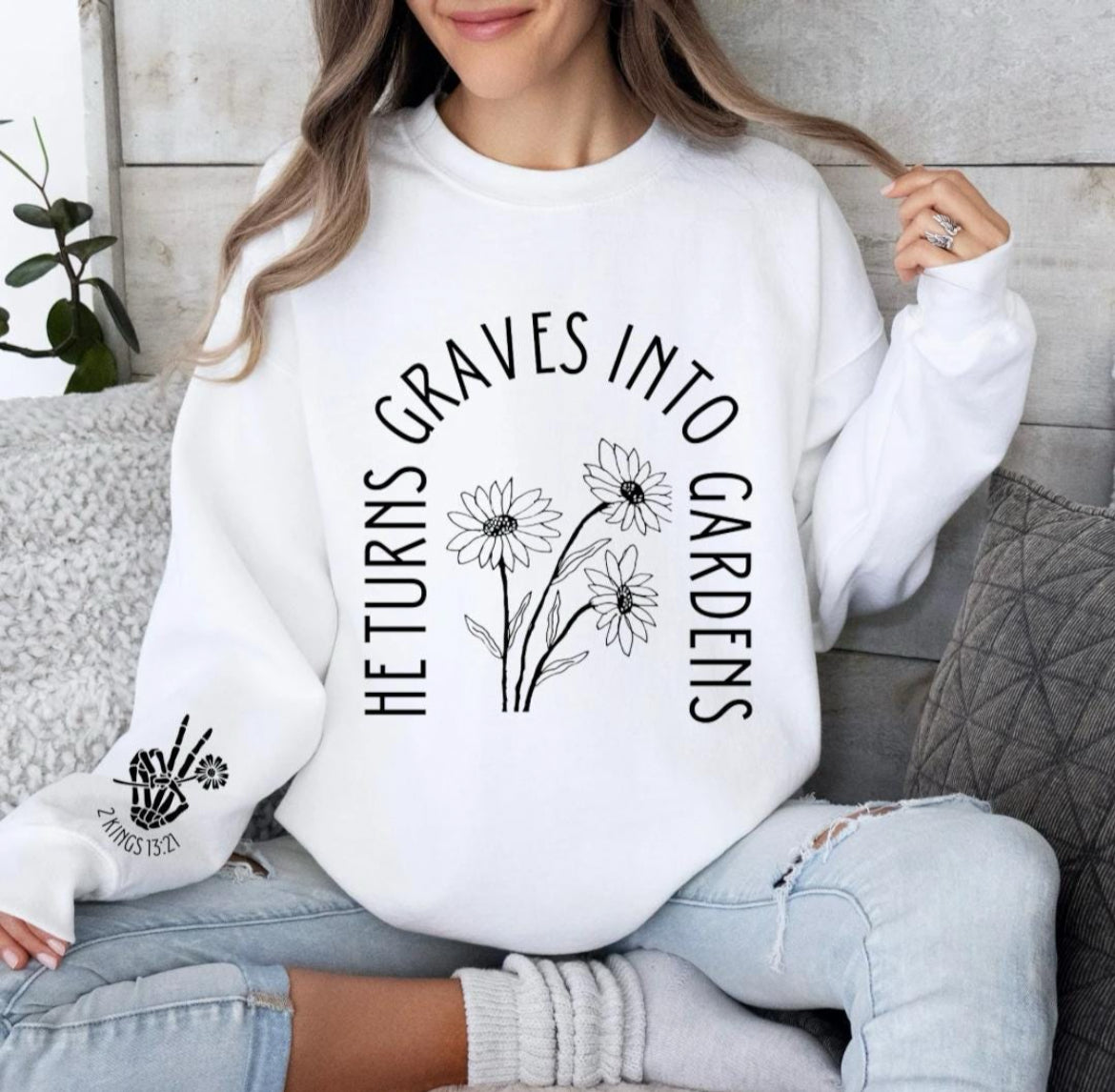 White crewneck sweatshirt that says, "He turn graves into gardens." The words are in the shape of a grave. In the middle are flowers. On the sleeve is a peace sign bones hand holding a flower and underneath it says, "2 Kings 13 21."