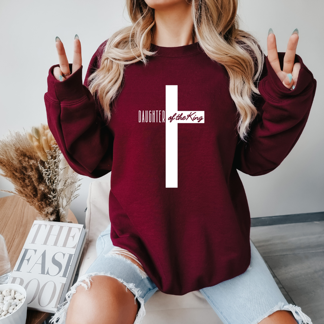 maroon crewneck sweatshirt that says, "daughter of the King" across the middle of a large cross. 