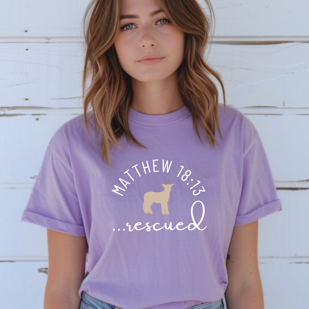 lavender crewneck T-Shirt that says, "He left the 99 to rescue me" with a large tan cross. On the front it says, "Matthew 18:13 rescued" with a tan sheep. 