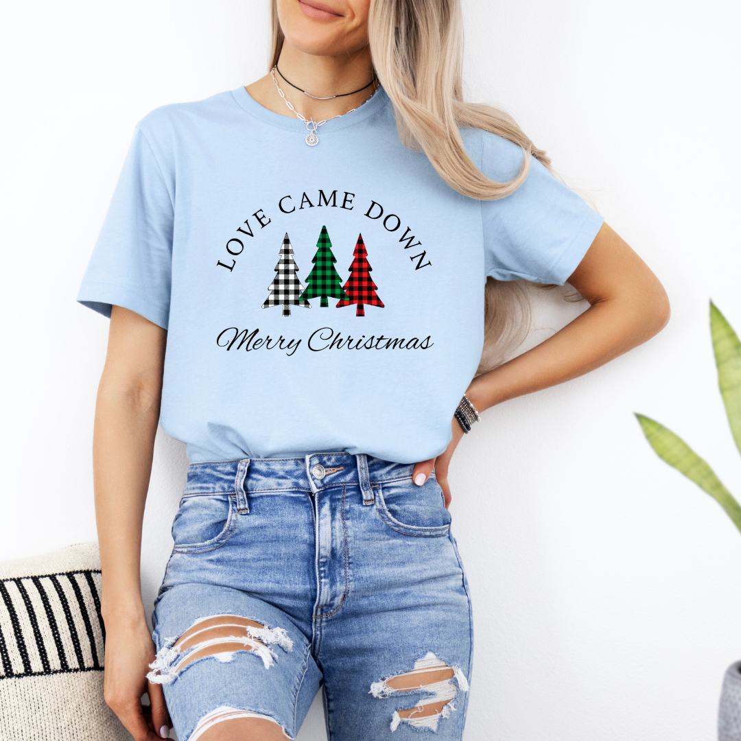 Baby blue crewneck t-shirt that says, "Love came down. Merry Christmas." It features 3 Christmas trees in buffalo plaid green, red and white.