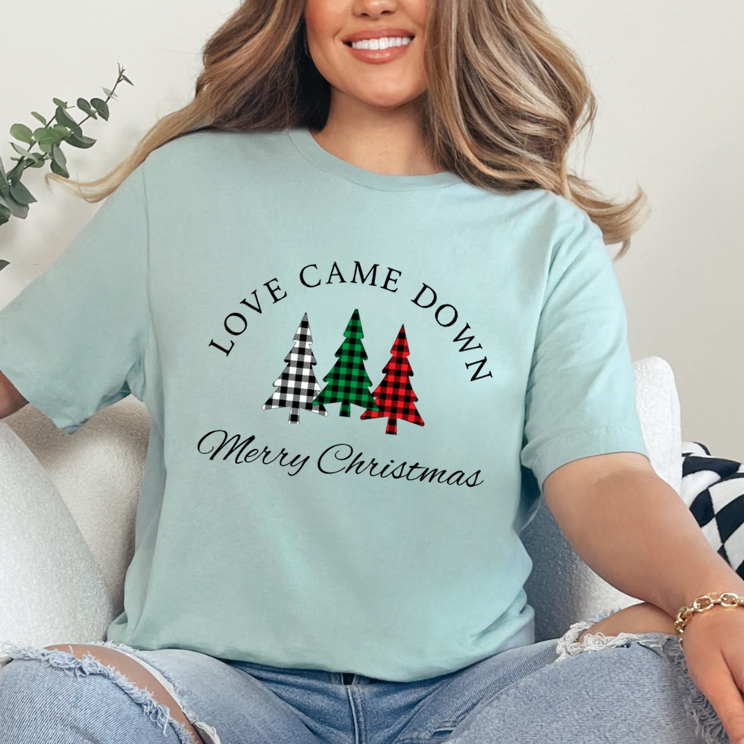 Dusty green crewneck t-shirt that says, "Love came down. Merry Christmas." It features 3 Christmas trees in buffalo plaid green, red and white.