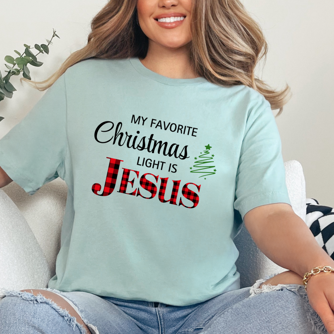 Dusty green crewneck t-shirt that says, "My favorite Christmas light is Jesus." Jesus is in red buffalo plaid, there is a Christmas tree graphic.