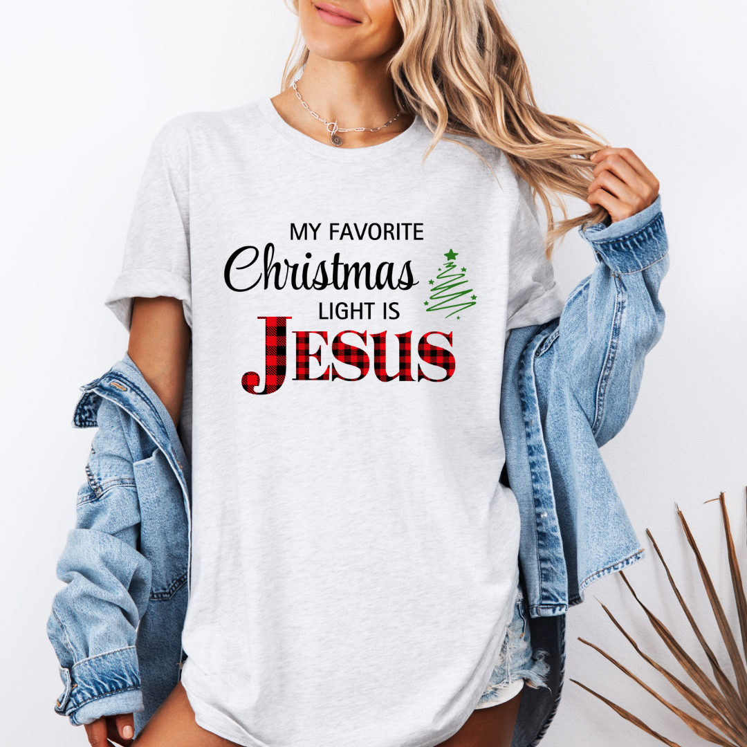 Ash white crewneck t-shirt that says, "My favorite Christmas light is Jesus." Jesus is in red buffalo plaid, there is a Christmas tree graphic.