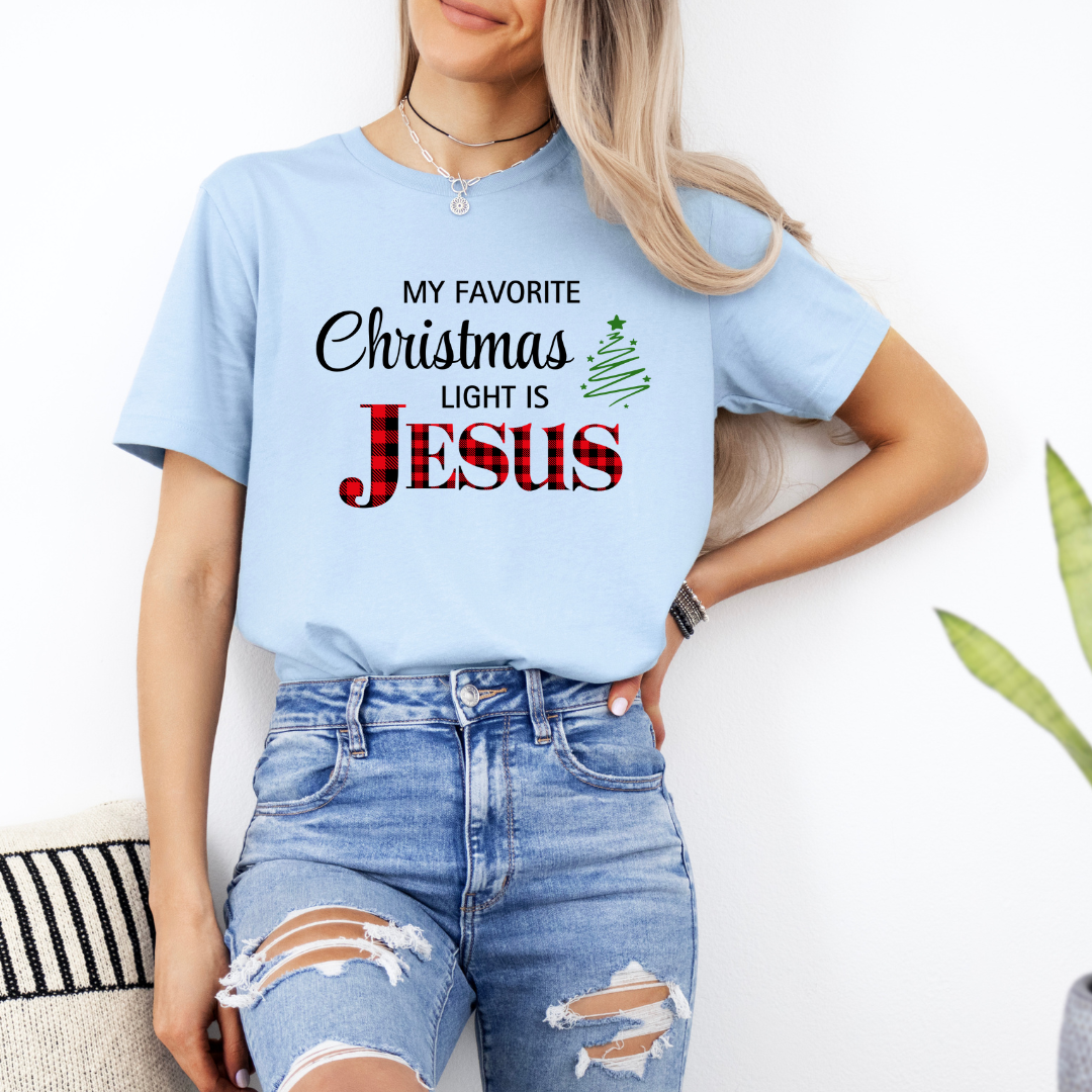 Baby blue crewneck t-shirt that says, "My favorite Christmas light is Jesus." Jesus is in red buffalo plaid, there is a Christmas tree graphic.